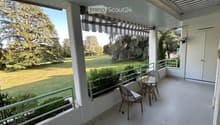 Balcony with white tiled floor, green metal railing, striped awning, small round glass table, and two wicker chairs. Offers a view of a green lawn and trees.