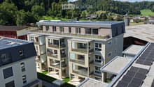 Modern multi-story residential building with solar panels on the roof, surrounded by greenery and a scenic landscape.