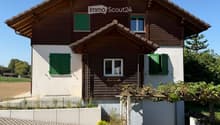 Two-story house with wooden siding, green shutters, and a raised ground floor. Features a garage and small garden area.