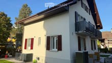 Two-story house with white walls, red shutters, and a balcony on the upper floor. The house has a well-maintained garden with a barbecue, a small fence, and outdoor furniture.