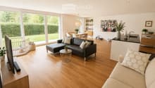 Open-plan living and dining area with wooden flooring, large windows, a black leather sofa, a beige sofa, a coffee table, a dining table with chairs, a kitchen counter, and various decorative items.