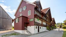 A two-story building with red brick walls and green shutters, featuring a wooden balcony and a sign reading "Mietshaus zur Gögenburg".