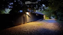 A modern, single-story building with a dark wooden facade and a gravel driveway, illuminated by warm lighting at dusk.