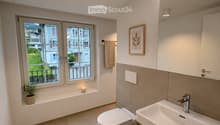 Modern bathroom with a toilet, sink, and a window offering a view of the outside. Includes a towel, a small plant, a picture frame, and a rug on the floor.