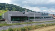 Modern, single-story building with a grey metal facade and large glass windows, situated near a road with a landscaped area and distant hills.