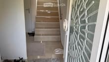 Hallway with a staircase leading upwards, a glass door with decorative pattern on the right, and a gray rug on the floor. Shoes and slippers are placed near the entrance.