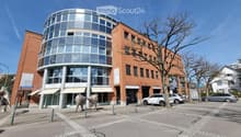 Modern multi-story building with a mix of red brick and glass facade, featuring a rounded glass tower section and several parked cars in front.