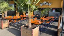 Outdoor seating area with wooden tables and chairs arranged in rows, featuring multiple potted palm trees on the ground, yellow building, signage with the text 'Dortbeizl', and an outdoor seating arrangement for customers.