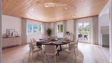 A modern dining room with a round wooden dining table surrounded by six chairs, set with plates, cutlery, and glasses. The room features light wooden flooring, white walls, large windows with curtains, and a sleek cabinet with decorative items. A potted p