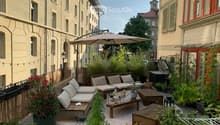 Cozy rooftop terrace with wooden seating, cushions, small table, potted plants, and an umbrella, overlooking a cityscape with historic buildings.