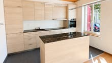 Modern kitchen with light wooden cabinets, dark granite countertop, and a central island. Features include a sink, oven, and pendant lighting.