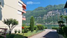 A multi-story residential building with balconies overlooking a scenic lake and mountain backdrop. The area includes greenery, trees, and a paved pathway.