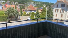 Balcony with a view of a town, featuring tiled flooring and a metal railing, overlooking a street with parked cars and surrounding buildings.