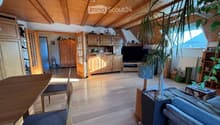 Bright living room with wooden floors, exposed wooden beams, large windows, a sofa, a coffee table, a dining table with chairs, a television, and a large potted plant.