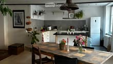 Open-plan kitchen and dining area with wooden flooring, a large wooden dining table with chairs, pendant lighting, and various kitchen appliances and decor.