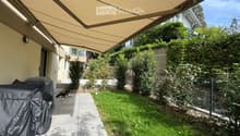 A well-maintained garden area with a covered patio, a covered grill, a small dog, and a variety of plants and shrubs.