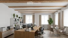 Modern office space with exposed wooden beams, large windows, office desk with multiple monitors, chairs, laptop, printer, bookshelf with books and plants, and a small round glass table with chairs.