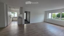 Empty room with wooden flooring, white walls, large windows, and a small kitchen area visible in the background.