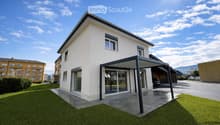 Modern two-story house with white exterior, large windows, and a covered patio area. The house features a well-maintained lawn and is situated in a residential area with neighboring buildings visible in the background.