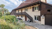 Two-story house with a tiled roof, wooden balcony, and stone facade. Features include multiple windows with shutters, a wooden staircase leading to the entrance, and a small garden area with plants and a bench.