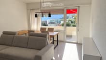 Modern living and dining area with a grey sectional sofa, wooden dining table, chairs, pendant lighting, and large windows offering a scenic view.
