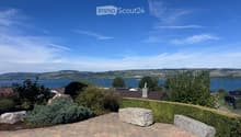 Garden with a view of a lake and surrounding hills, featuring a paved patio, stone benches, and various shrubs and trees.
