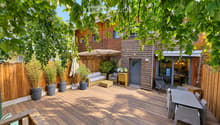 Wooden decked outdoor area with a sofa, coffee table, dining table, chairs, and potted plants, surrounded by wooden fencing and trees.