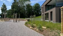 Modern two-story house with dark brown siding and large windows, surrounded by a gravel pathway and greenery.