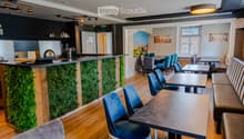 Modern restaurant interior with wooden flooring, blue chairs, tables, a bar counter with a green moss wall, and a seating area with a sofa and bench.
