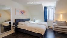 Cozy bedroom with two single beds, a sofa, small side tables with lamps, a large mirror, and a colorful painting on the wall. The room features dark wooden flooring and large windows with blue curtains.