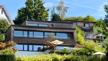 Modern two-story house with large windows and wooden exterior, surrounded by lush greenery and trees.