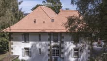 Two-story house with a red-tiled roof, white walls, multiple windows with shutters, and a small staircase leading to the entrance.