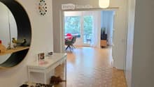 Modern hallway with wooden flooring, white walls, a hanging light fixture, and a round mirror on the wall. A small table with decorative items and a bench are visible, leading to an open area with chairs and a table.