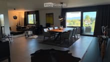 Modern living and dining area with large windows offering a scenic view, featuring a sofa, coffee table, ottoman, dining table with chairs, and decorative vases.