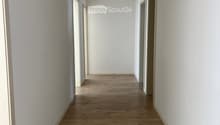 Empty hallway with light wooden flooring and white walls, featuring several doors and ceiling lights.