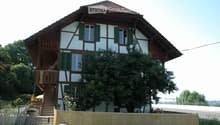 Two-story traditional house with white and brown timber framing, green shutters, and a wooden balcony accessible via stairs.