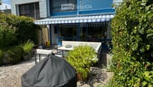 Outdoor patio area with a covered seating area, featuring a sofa, table, chairs, and a covered grill, surrounded by plants and a modern house with blue shutters.