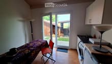 A small, furnished room with a wooden floor, a covered table with a chair, a washing machine, a sink, and a floor lamp. A sliding glass door leads to a backyard.