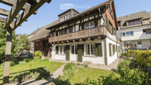Traditional two-story house with white walls, green shutters, and a wooden balcony. The house features a well-maintained garden with a paved pathway leading to the entrance.