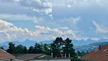 Residential buildings with tiled roofs, surrounded by trees and distant mountains under a partly cloudy sky.