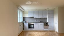 Empty kitchen with light grey cabinets, wooden floor, and a window on the left side.