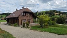A two-story wooden house with a brown tiled roof, multiple windows, and a chimney, situated in a rural landscape with green hills and trees.