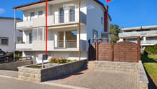 Two-story modern house with white and gray exterior, large windows, balconies, and a wooden gate entrance.