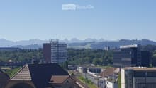 A panoramic view of a cityscape with a mix of modern and traditional buildings, surrounded by green fields and distant mountains under a clear blue sky.