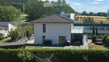 A modern two-story house with a tiled roof, surrounded by a well-maintained lawn, hedges, and a gravel driveway. The house features large windows and a balcony area with outdoor furniture.