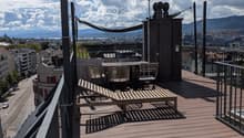 Wooden deck terrace with a table, chairs, and a lounge chair, overlooking a cityscape with mountains in the background.