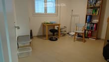 A small, furnished room with a tiled floor, a wooden table with a chair and stool, a bookshelf filled with books and binders, and several storage bins along the walls.
