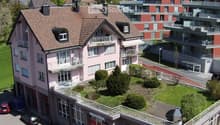Aerial view of a two-story residential building with a pink facade, multiple balconies, and a well-maintained garden area. The building features a garage and parking area, with neighboring structures visible in the background.
