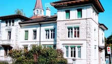 A large, white stone building with multiple stories, green shutters, and a prominent tower with a pointed roof. The building features a balcony and is surrounded by greenery and a fence.