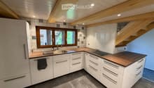 Modern kitchen with white cabinets, wooden countertops, stainless steel sink, and built-in stove. Features exposed wooden beams on the ceiling and large windows.